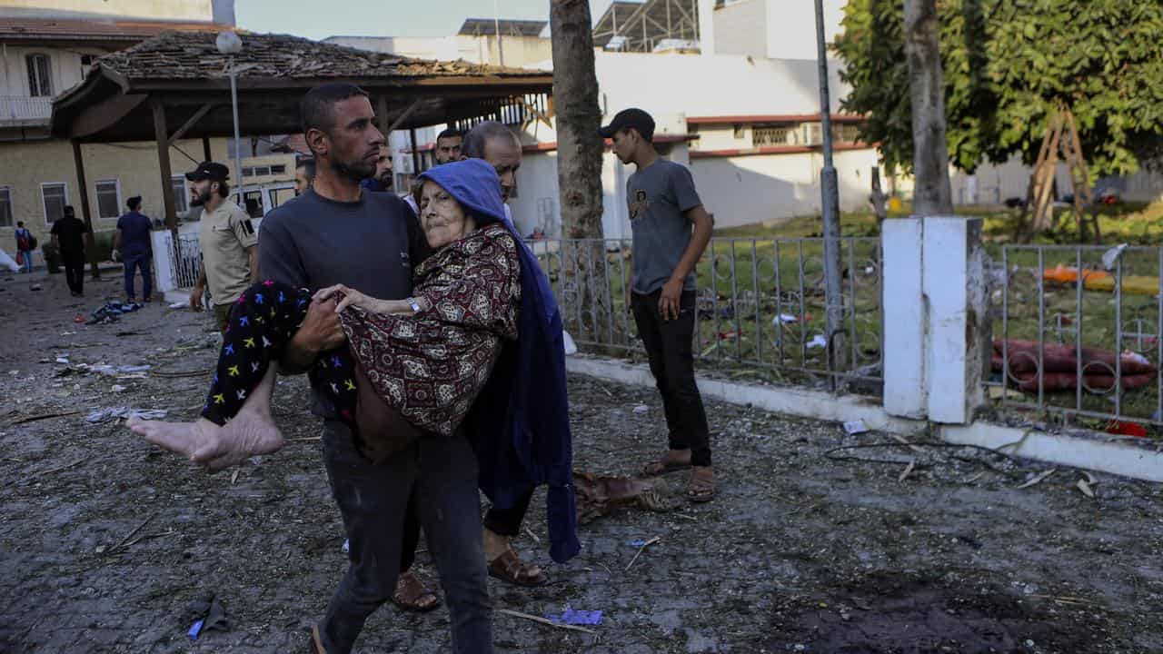 Man carries elderly woman past site of deadly Gaza hospital explosion.