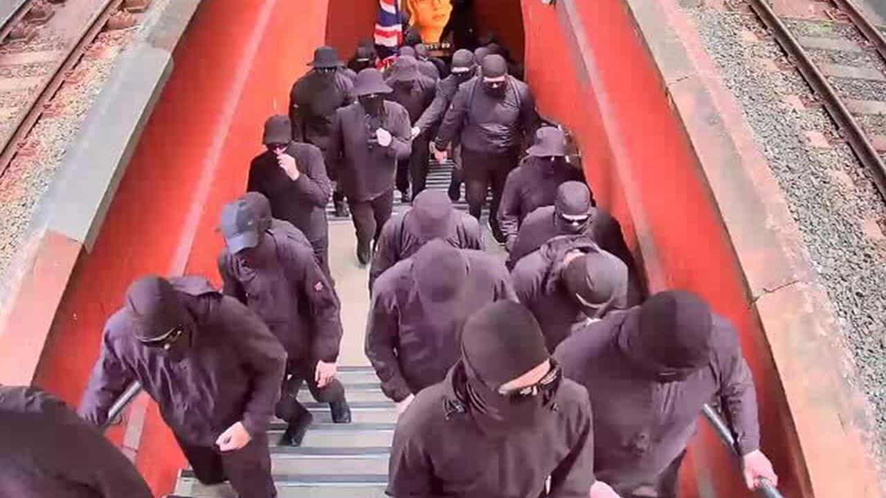 About 20 men all dressed in black walk up stairs to a train platform  